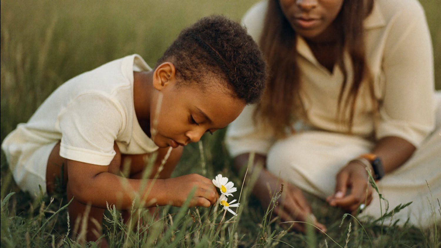 A young child crouched in tall grass, examining a wildflower, a parent softly out of focus behind