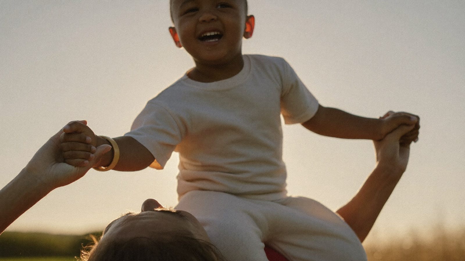 A laughing child rides on a parent's shoulders at golden hour, holding their hands, a soft band on one wrist